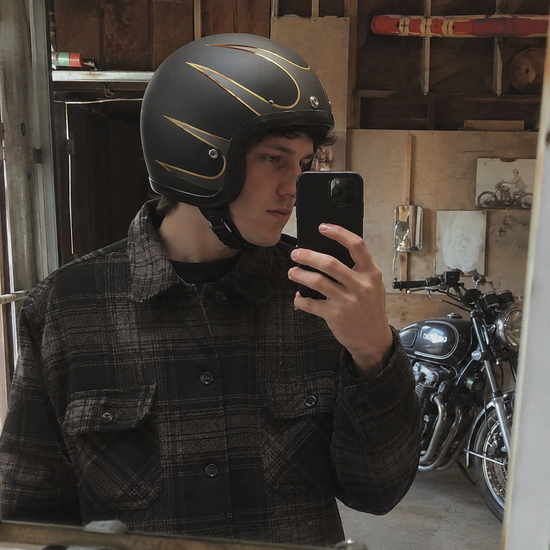 Young rider taking a mirror selfie in a garage, wearing a matte black open-face helmet with gold flames, a vintage motorcycle behind him