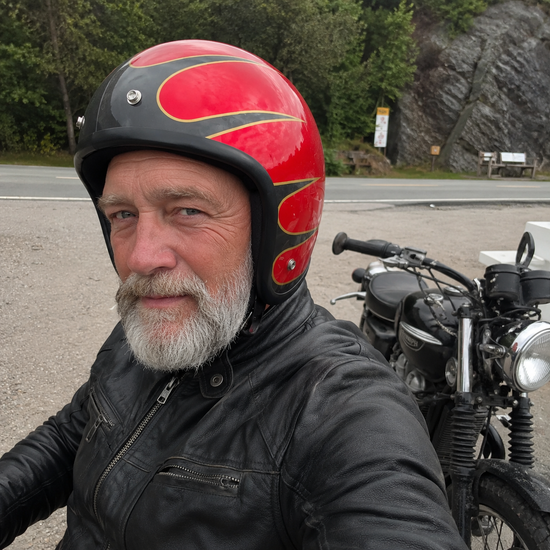 Bearded rider in a red flame open-face helmet and leather jacket, stopped beside his classic motorcycle on a mountain road